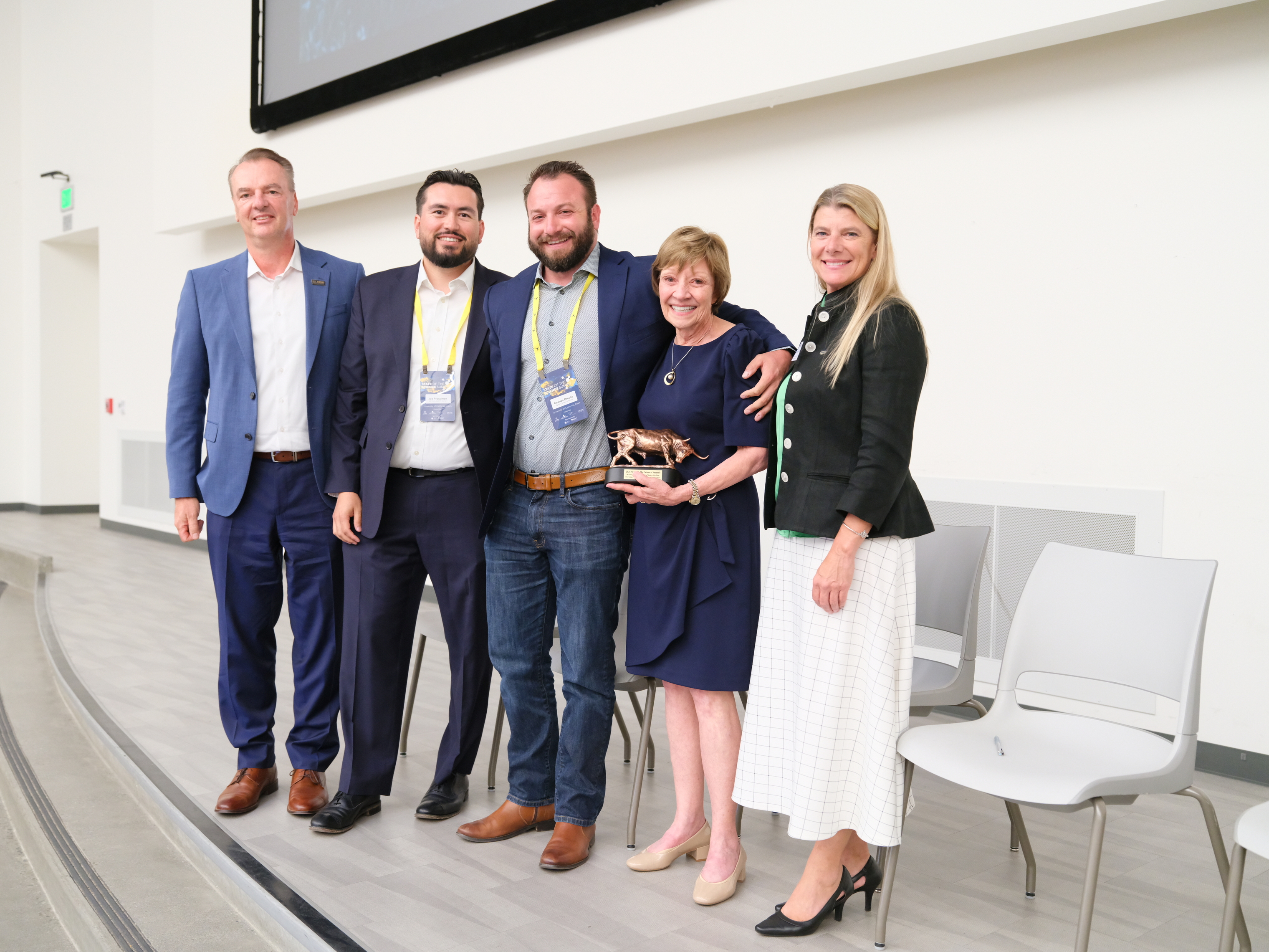 Group photo — six smiling professionals on stage; one woman holds a trophy