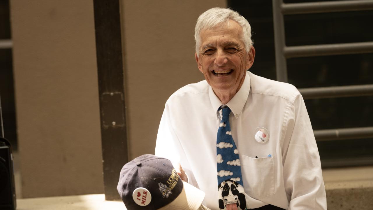 Smiling elderly man seated outdoors in white shirt and patterned tie, holding a cap