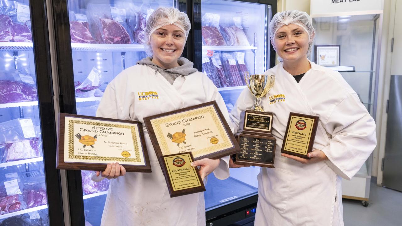Two smiling workers in white lab coats and hairnets holding awards in a meat cooler