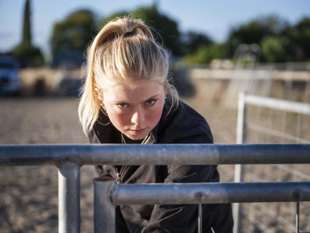 Teen girl with ponytail leaning on metal rail, intense gaze, sunlit outdoor court
