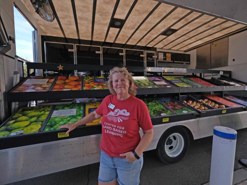 Smiling woman in red shirt stands in front of a mobile produce truck of fruit and veg