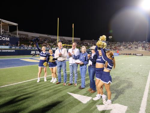 Night football stadium: group of people posed on turf, flanked by cheerleaders under bright lights