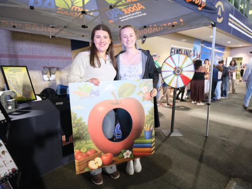 Two smiling women hold a large peach-shaped frame at an indoor market booth