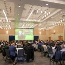 A ballroom is full of people sitting in chairs around round tables listening to a presentation. 